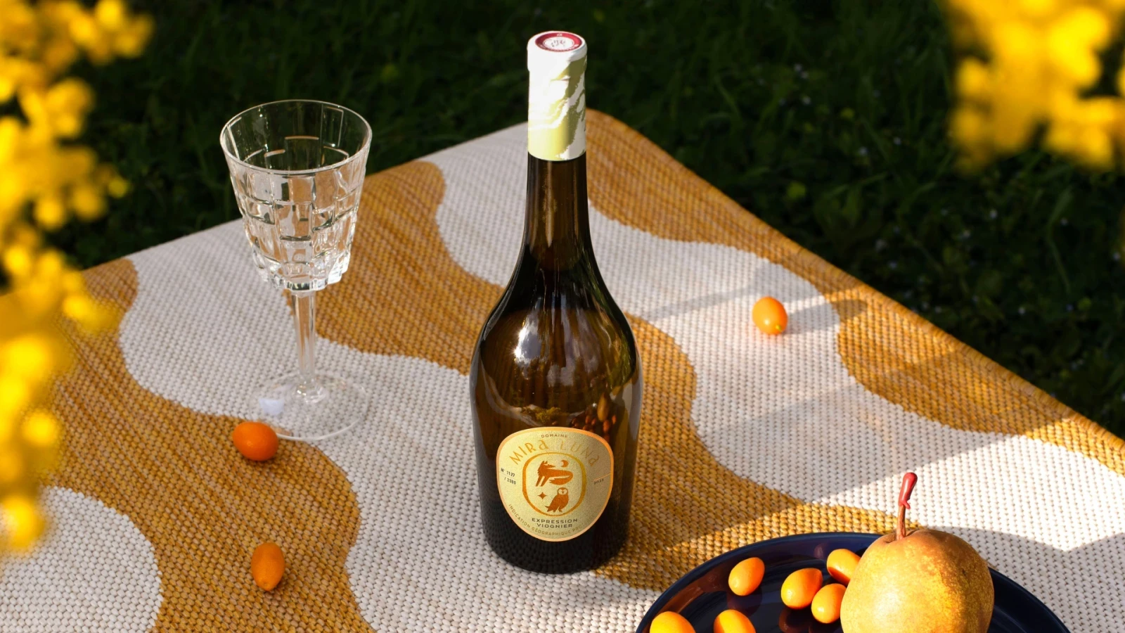 Photographie d'une bouteille de vin, un verre et un panier de fruit sur une belle nape colorée
